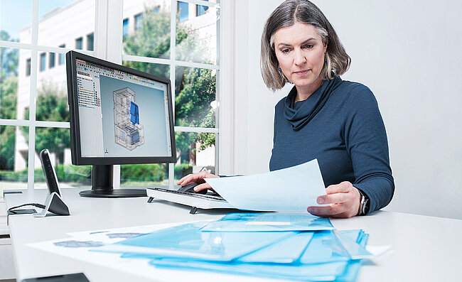 Eine Frau sitzt am Schreibtisch vor ihrem Computer mit technischen Zeichnungen, vor ihr liegen Dokumente